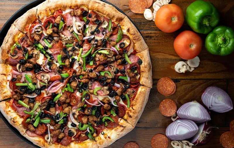 Fresh pizza on a wooden table surrounded by tomatoes, peppers, and onions