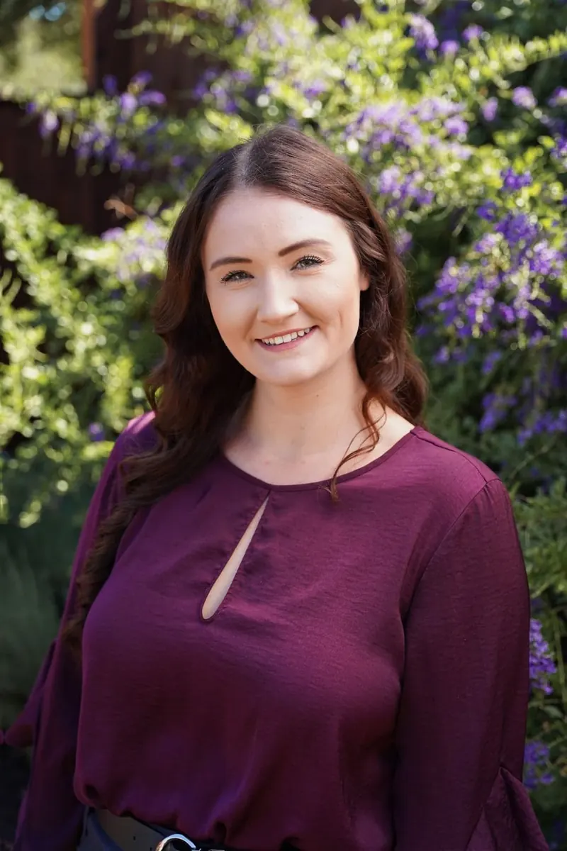 Smiling woman with long auburn hair in a plum top in front of purple flowers